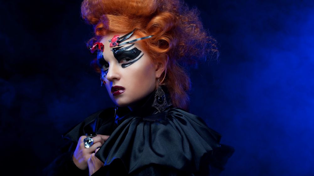 Künstlerisches Porträt einer Frau mit voluminösen roten Haaren, kreativem Make-up und einem dramatischen Cape