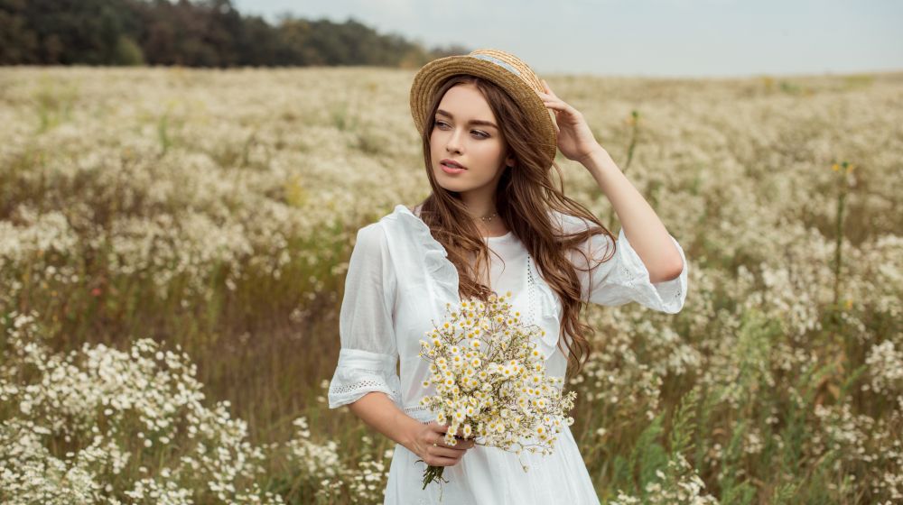 Junge Frau in weißem Kleid mit Hut und Blumen in der Hand auf einer Wiese, nach professionellem Styling